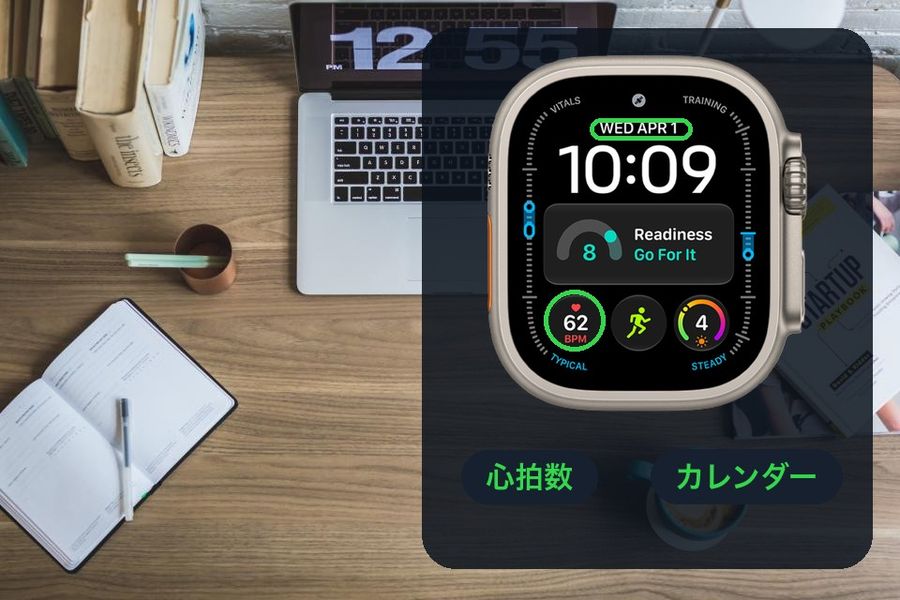 A desk photo with a dark panel showing Apple's Modular Ultra watch face, with two of its complications named: カレンダー (the date) and 心拍数 (heart rate)