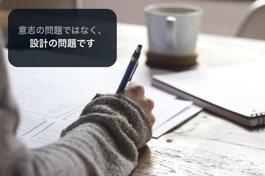 A close-up of a hand writing, with a dark panel reading 意志の問題ではなく、設計の問題です (it's not a matter of willpower, it's a matter of design)