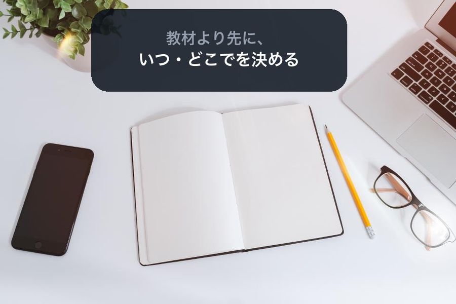 A flat-lay desk with a blank notebook, phone and glasses, with a dark panel reading 教材より先に、いつ・どこでを決める (decide when and where before the materials)