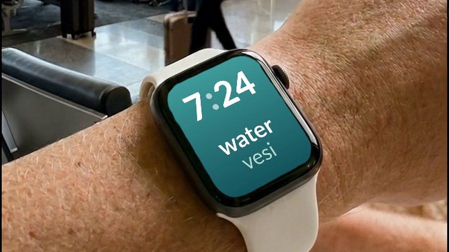 Apple Watch ranteessa lentokentällä, näytöllä englannin sana water ja sen suomennos vesi kellonajan alla