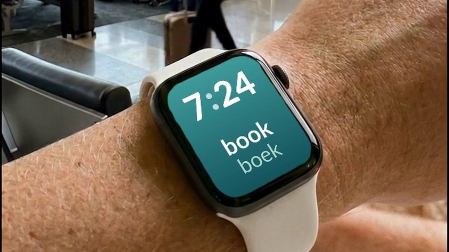 Een Apple Watch om een pols op een luchthaven, met het woord book en de vertaling boek eronder, naast de tijd