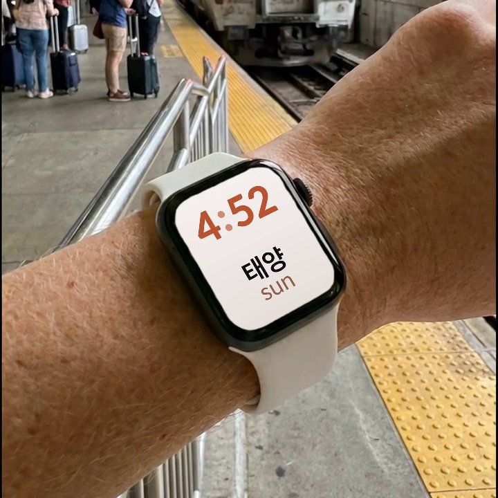 An Apple Watch on a wrist at a train station, showing the Korean word 태양 and its English translation, sun